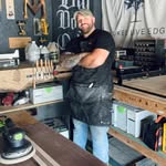 Man standing in a workshop wearing a black apron and hat with wood tools and materials around him.