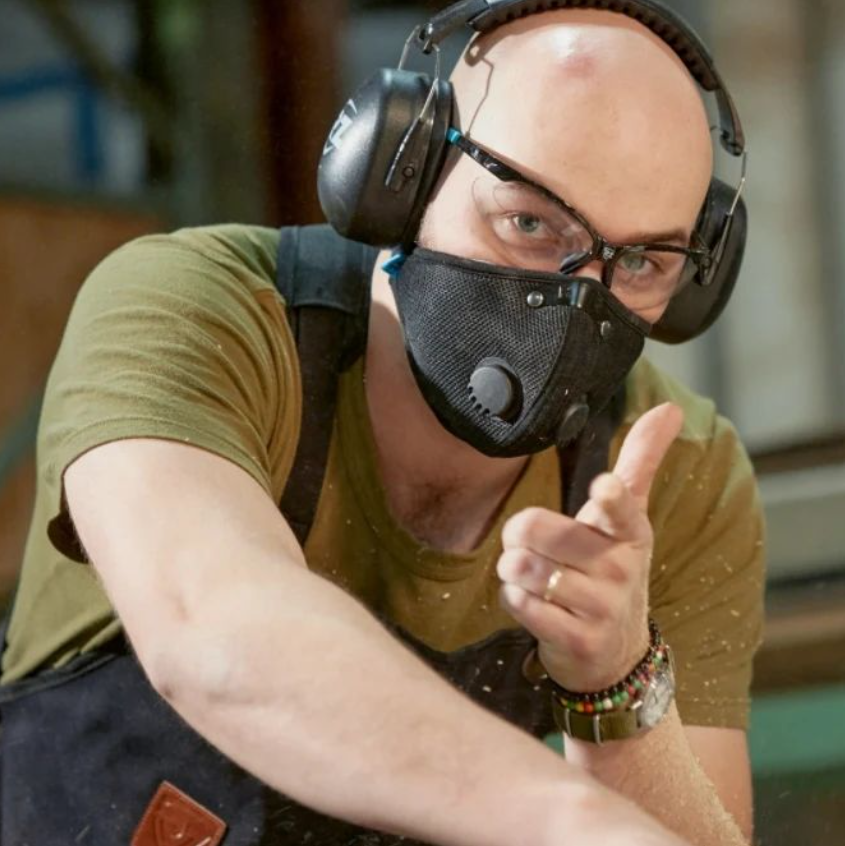 Man wearing a black respirator mask with earmuff hearing protection and safety glasses while pointing playfully.