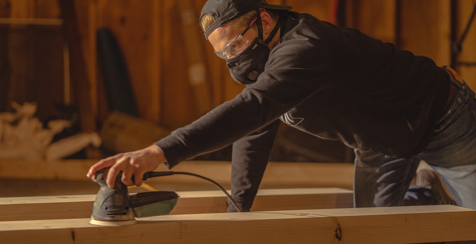 Man on a jobsite wearing an rz mask m2.5 mask while using a palm sander on a large beam. he is also wearing a hat, safety glasses and a sweatshirt