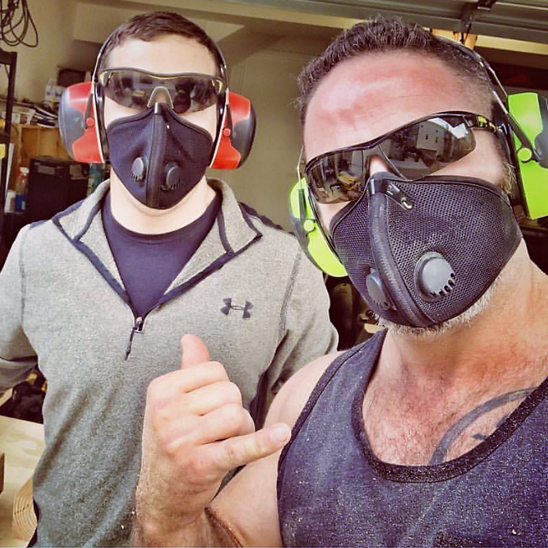 Two men wearing respirator masks, safety glasses, and earmuff hearing protection posing in a workshop.