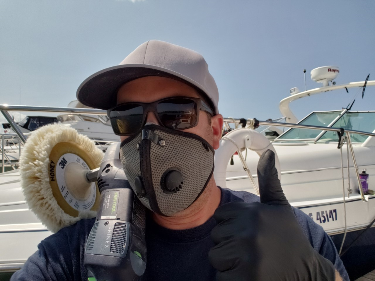 Man wearing a gray respirator mask and sunglasses giving a thumbs up while polishing a boat at a marina.