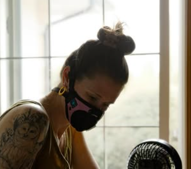 Woman wearing a pink respirator mask while working indoors near a small fan.