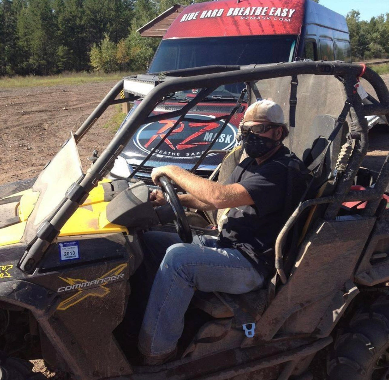 Man in a UTV wearing an RZ Mask M1 Black mask to protect from dust while on the trail. There is a van with the RZ Mask logo in the background.