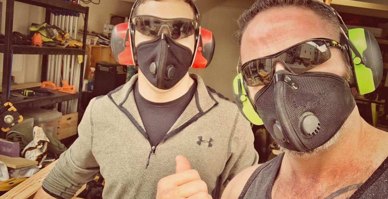 Two men wearing respirator masks, safety glasses, and earmuff hearing protection posing together in a workshop.