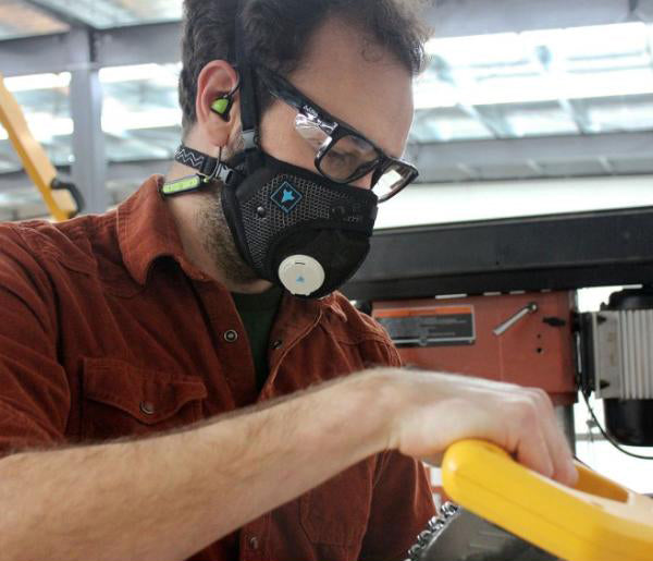 Man wearing a black respirator mask, safety glasses, and earplugs while operating equipment in a workshop.