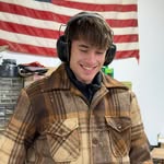 Man wearing earmuff hearing protection and a brown plaid jacket smiling in a workshop with an American flag behind him.