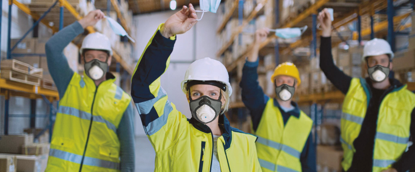 Workers in high visibility vests and hard hats wearing respirator masks while holding disposable masks in a warehouse.