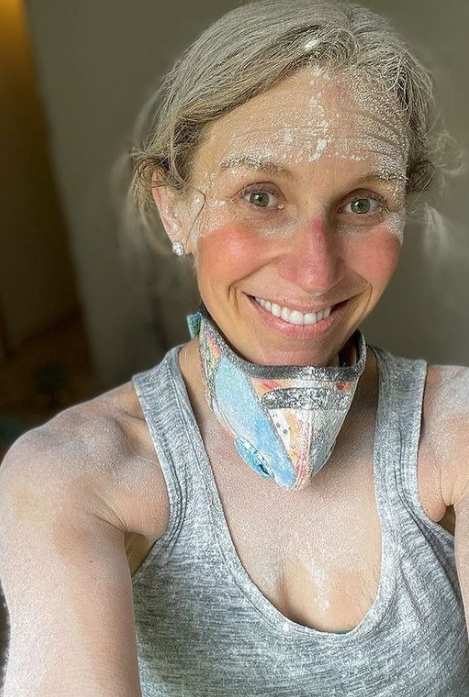Woman covered in sanding dust smiling with a colorful respirator mask pulled down around her neck.