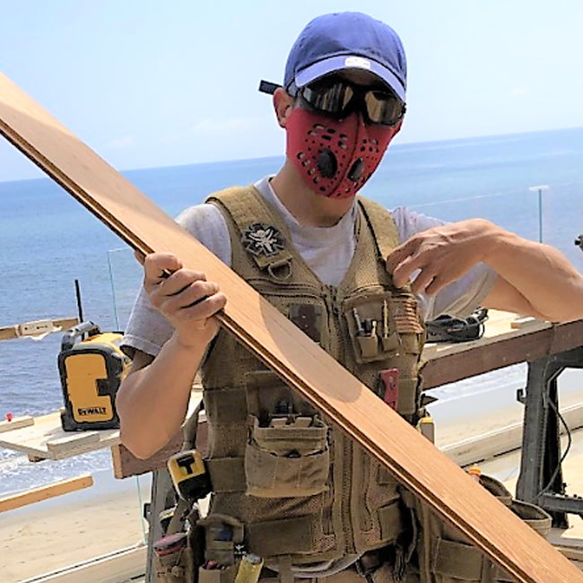 Man on a construction site holding a piece of wood while wearing an rz mask m1 red for protection against dust and particulates. he is also wearing a blue hat and safety glasses. 