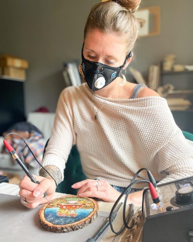 Woman woodburning with a RZM3 mask on, surrounded by art supplies.