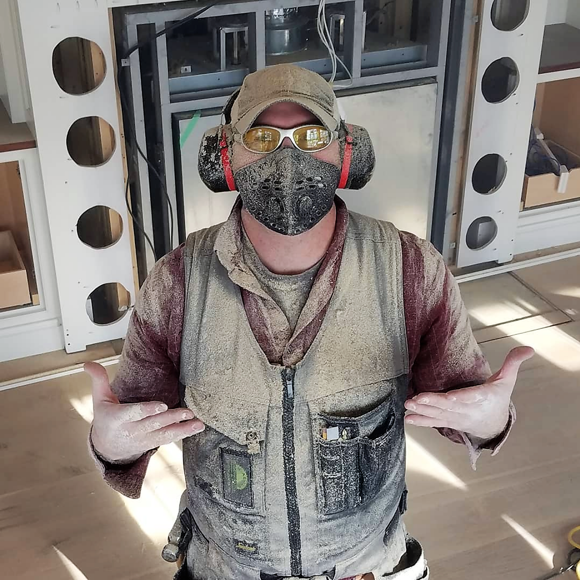 Worker covered in heavy sanding dust wearing a respirator mask, earmuff hearing protection, and safety glasses.