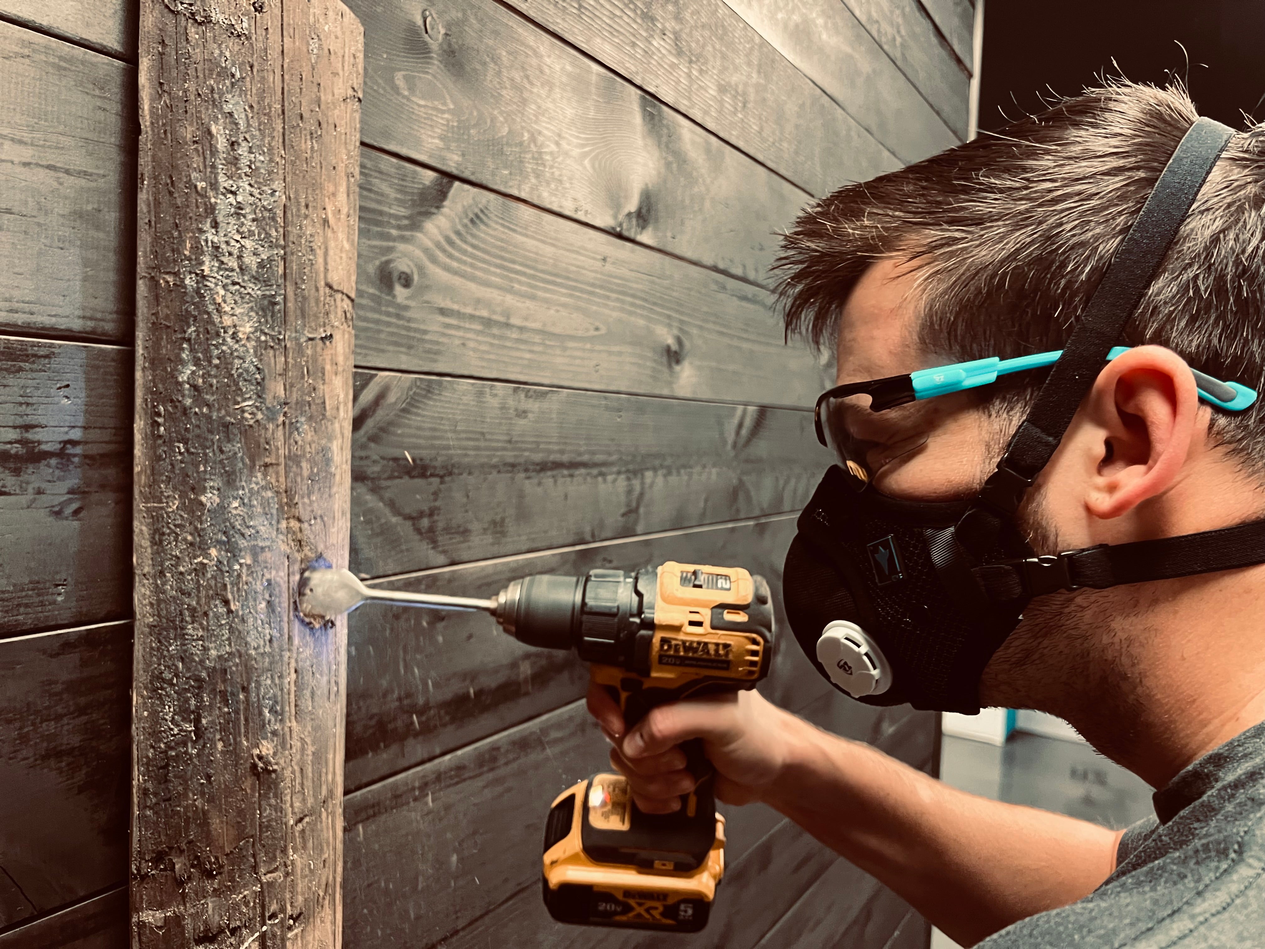 Man wearing a black respirator mask and safety glasses using a drill on a wooden wall.