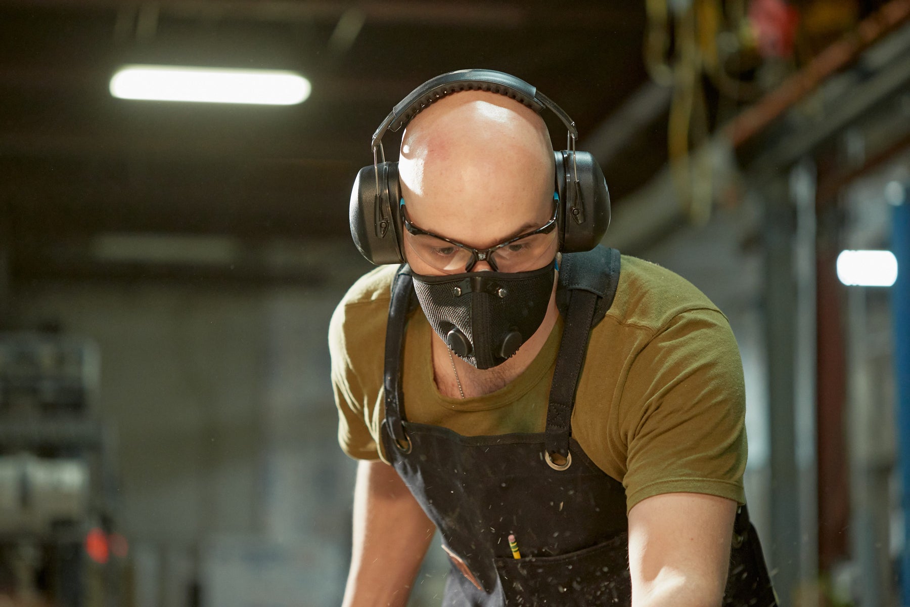 A woodworker stands in a wood shop wearing an RZ Mask M2 Black Dust Mask for protection against dust, sawdust and other particulates while working on projects. He also wears RZ Earmuffs and Safety Glasses, along with an RZ Apron.