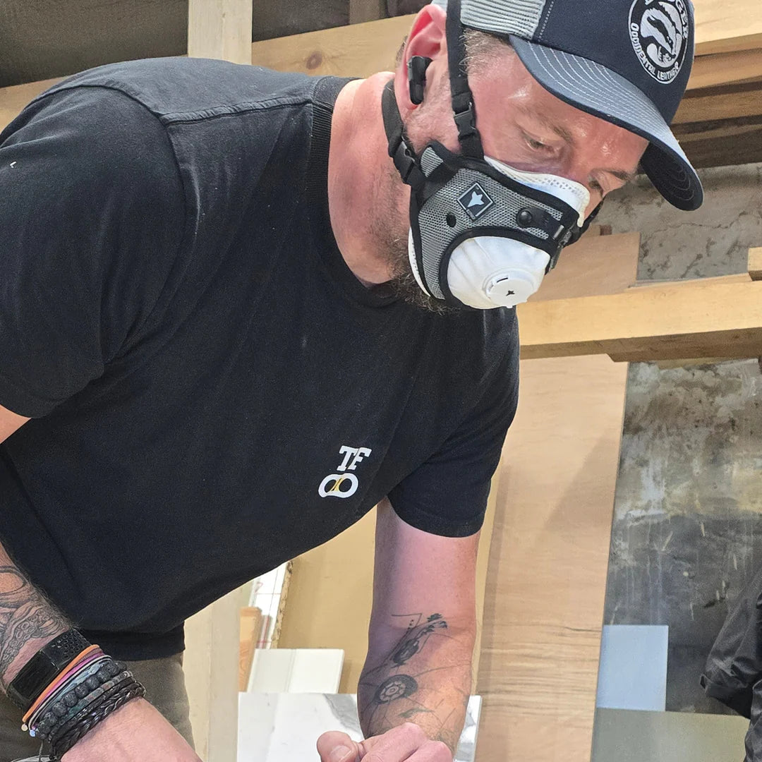 Man wearing a black shirt, cap, and dust mask working on a wooden surface.