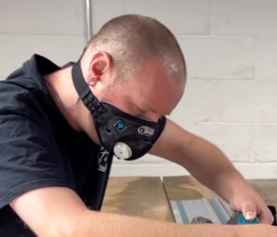 Man wearing a black respirator mask while sanding or working on a project in a workshop.