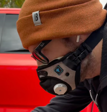 Man wearing a tan respirator mask and beanie while working beside a red vehicle.