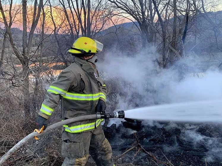 Firefighter in full gear spraying water on smoky brush during a small wildfire containment.